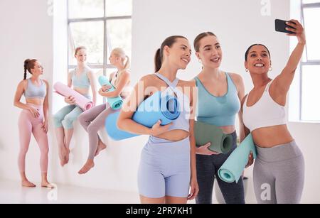 Happy friends taking a selfie before yoga class. Group of women taking a photo on a cellphone before pilates class. Young women bonding before an exercise class. Women using cellphone in yoga class Stock Photo