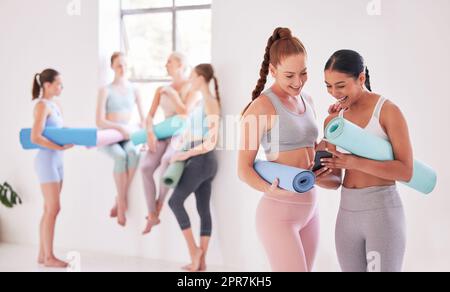 Friends looking at an online app on a cellphone. Friends bonding before yoga class. Women using a smartphone before pilates exercise. Cheerful friends talking before yoga class in the studio Stock Photo