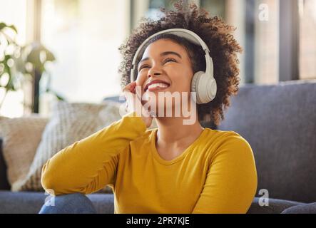 Young cheerful mixed race woman thinking while wearing headphones and listening to music at home. One content hispanic female with a curly afro enjoying music and daydreaming while sitting on the floor at home Stock Photo