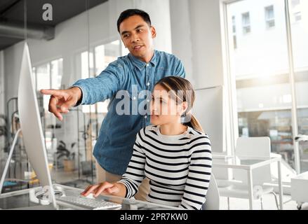 Two businesspeople working on a desktop computer together at work. Young asian male manager training a new employee in an office. Female businesswoman getting help from her boss at work Stock Photo