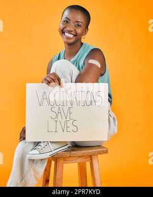 African american covid vaccinated woman showing plaster on arm, holding poster. Portrait of smiling black woman isolated against yellow studio background with copyspace. Promote corona vaccine on sign Stock Photo