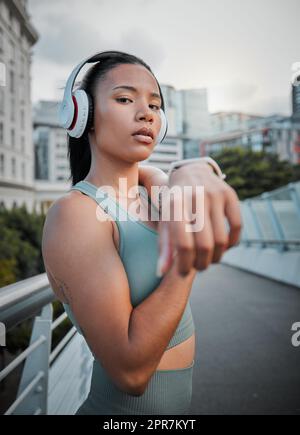 Young mixed race female athlete wearing headphones and listening to music while stretching her muscles before a run outside in the city. Warming up before exercising to improve her health and lifestyle Stock Photo