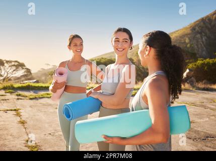 Beautiful yoga women bonding and holding yoga mats in outdoor practice in remote nature. Diverse group of young smiling active friends standing together. Three happy people getting ready to be mindful Stock Photo