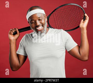 One happy mature african american man standing against a red background in studio and posing with a tennis racquet. Smiling black man feeling fit and sporty while playing a match. Ready for the court Stock Photo