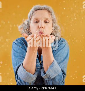 One happy mature caucasian woman blowing confetti out of the palm of her hands while posing against a yellow background in the studio. Smiling white lady showing joy and happiness while celebrating Stock Photo