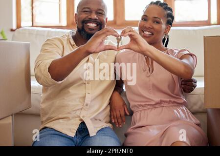 A couple holding keys, making a heart sign with their hands at home. Portrait of a smiling, happy african american man and woman in love, sitting in the lounge of their new home, looking at the camera Stock Photo