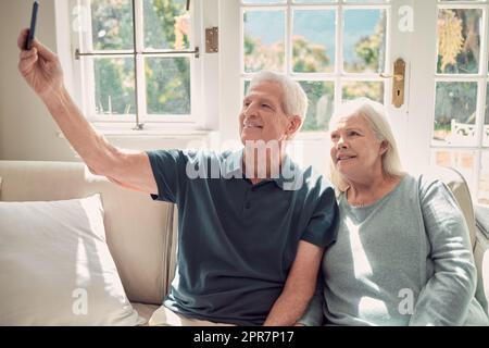 Lets see what this selfie fuss is all about. Cropped shot of an affectionate senior couple sitting in their living room at home. Stock Photo