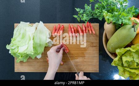 Closeup hand holding knife slices red tomatoes into pieces on a wooden chopping board. Top View. A wooden bowl with a variety of fruits and vegetables is placed on the kitchen counter. Stock Photo