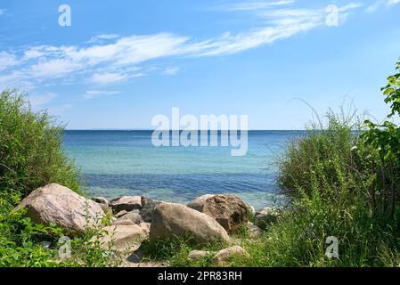 A shallow rocky coast on a calm quiet beach day during summer with grass growing on the shore. Scenic view of a crystal blue ocean with clear blue skies and white clouds during a warm summer day Stock Photo