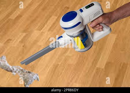 The male hand holds hold a modern cordless battery-powered vacuum cleaner over abstract blurred background. Space. Household cleanliness and hygiene. Stock Photo