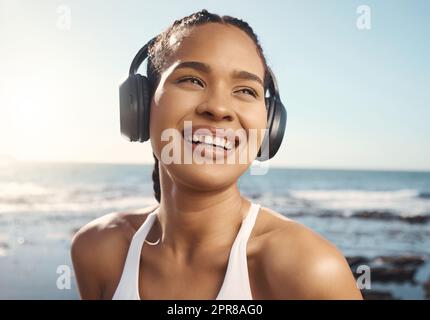 Young mixed race female athlete looking happy and smiling while wearing headphones and listening to music while out for a run along the promenade. Exercise for a healthier mind and body Stock Photo