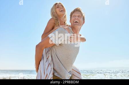 A young loving couple having fun with a piggyback ride, enjoying a day at the beach while smiling hugging and showing at the ocean. Romantic man carrying his wife on his back while on vacation Stock Photo