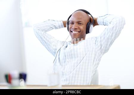 Taking a breather at work. a relaxed-looking african american designer leaning back in his chair in front of a desktop computer in the office and wearing a headphones. Stock Photo