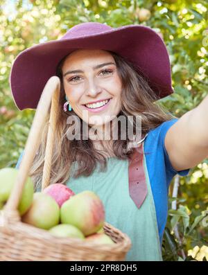 Cheerful farmer harvesting juicy organic fruit in season to eat. Portrait of a happy woman taking selfies while holding basket of fresh picked apples on sustainable orchard farm outside on sunny day Stock Photo