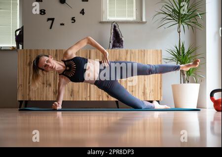 Young woman in sportswear working out at home taking care of her health and fitness. Making a side plank exercise for a strong core. On a mat in domes Stock Photo