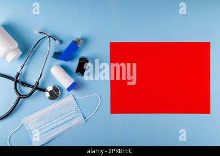 Top view of medical mask, stethoscope, oximeter, bottle of pills and inhalers on pastel blue background with bright red rectangular sheet of paper for Stock Photo