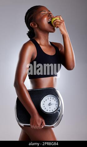 Healthy eating and regular exercise can help you lose weight safely. Studio shot of a sporty young woman eating an apple while holding a scale against a grey background. Stock Photo