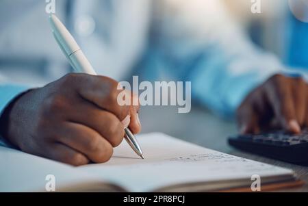 Closeup hands of african man writing in his diary while using a calculator. African american business man calculating figures while working late at night in his office. Putting in overtime after hours Stock Photo