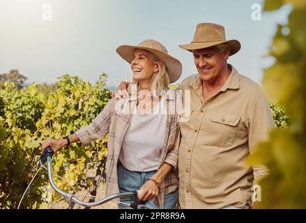 Smiling senior couple walking together while bonding on vineyard. Happy caucasian husband and wife pushing bicycle while enjoying day on farm after wine tasting on weekend. Man and woman on wine farm Stock Photo