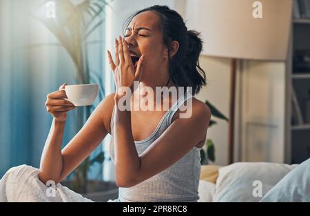A young mixed race woman yawning while lying in bed and drinking coffee. An attractive Hispanic female waking up from a her sleep and getting ready to begin her day Stock Photo