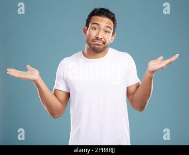 Handsome young mixed race man shrugging his shoulders while standing in studio isolated against a blue background. Confused hispanic male looking lost or clueless and making a, So what gesture Stock Photo