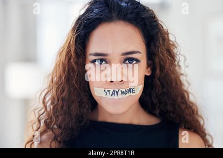 Its time to call out all forms of oppression and injustice. Portrait of a young woman with tape on her mouth that has the words stay woke written on it. Stock Photo