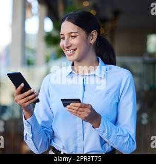 Young mixed race happy businesswoman using a credit card and phone to shop online at work. One hispanic woman smiling while paying for a purchase using her phone standing outside in the city Stock Photo
