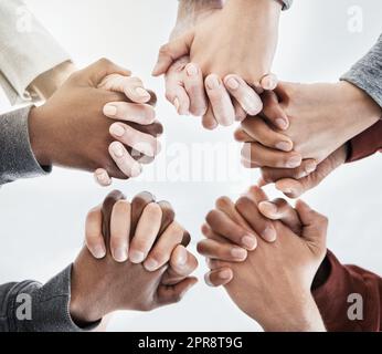 Below close up view of people holding hands in circle shape. A group of people putting their hands together while standing in a huddle inside against a clear grey background. Anything is possible with teamwork Stock Photo