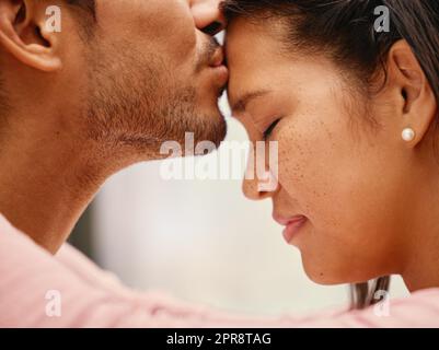 Closeup of mixed race man kissing his girlfriends forehead. Headshot of hispanic couple bonding and sharing an intimate moment at home. Beautiful woman with freckles feeling in love with boyfriend Stock Photo