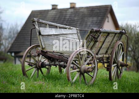 Old vintage rural rusty Amish timber open wagon. Horse/cow/ox-dragged vehicle for transporting hey, crops, or people. On grass in front of cottage. Stock Photo
