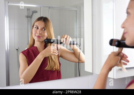 Close up of young woman latin using steam straightener to style hair in the mirror on bathroom Stock Photo