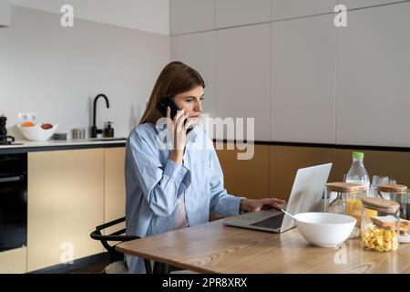 Young woman sitting at kitchen table using laptop computer and talking by phone with client Stock Photo
