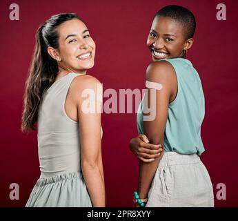 Covid vaccinated African american and mixed race women showing arm plaster. Portrait of two happy people isolated on red studio background with copyspace. Black woman and hispanic with corona vaccine Stock Photo