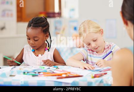 African american girl sitting at a table and colouring at pre-school or kindergarten with her caucasian classmate . Young female children using colourful pencils to draw pictures in a class at school Stock Photo