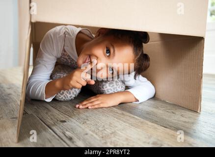 Portrait of a cute little hispanic girl playing with a cardboard box in a new apartment. Cute mixed race girl hiding in a box and smiling in a house. Relocation and moving day of big family Stock Photo