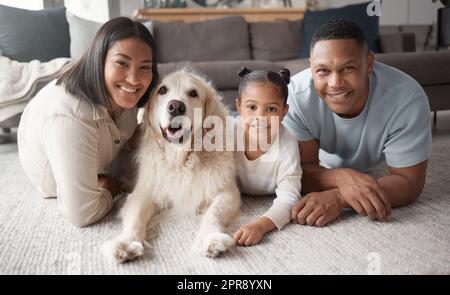 Portrait of a happy mixed race family of three relaxing on the lounge floor with their dog. Loving black family being affectionate with a foster animal. Young couple bonding with their daughter and rescued puppy at home Stock Photo