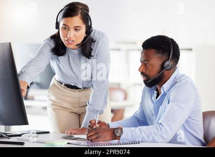 Young caucasian call centre telemarketing agent training new african american assistant on a computer in an office. Team leader troubleshooting solution with intern for customer service and sales support. Colleagues operating helpdesk together Stock Photo