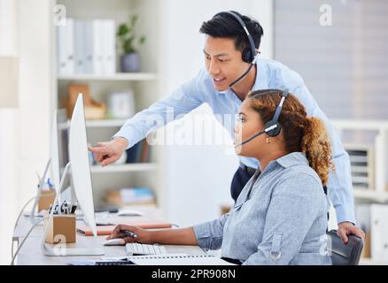 Young asian call centre telemarketing agent training new african american assistant on a computer in an office. Team leader troubleshooting solution with intern for customer service and sales support. Colleagues operating helpdesk together Stock Photo