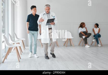 Lets go through to my office and well do a full assessment. a mature doctor meeting with a young man in the waiting room of a clinic. Stock Photo