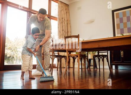 Mixed race father helping little boy sweep dust and dirt on wooden floor with broom for household chores at home. Cute boy helping dad with daily spring cleaning tasks. Kid learning to be responsible Stock Photo