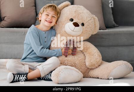 Portrait of one happy little caucasian boy smiling while hugging a big and cosy fluffy teddy bear on the floor in the lounge at home. Adorable kid relaxing and playing with soft stuffed toy alone Stock Photo
