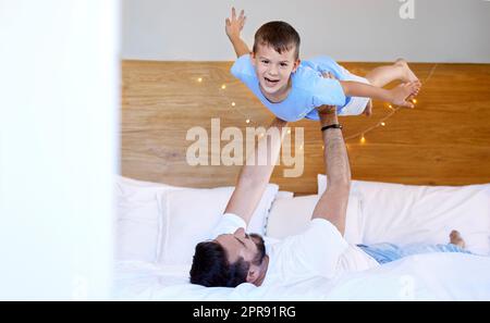 Caucasian father lifting his cute little son in the air to pretend to fly like a plane or superhero with arms out on a bed at home. Loving dad playing and bonding with cheerful kid in the morning Stock Photo