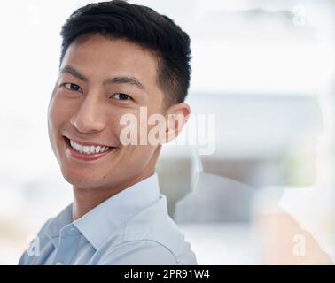 Portrait of one confident young asian business man and leader standing against office glass wall with copyspace. Face of happy successful male leader and entrepreneur showing motivation in his startup Stock Photo