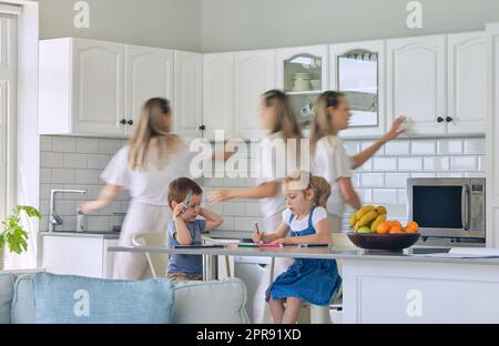 Busy mom feeding kids and cleaning the house. Little girl and boy sitting and doing homework while mother does chores. Young single parent having a nervous breakdown and looking overwhelmed Stock Photo