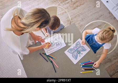 Above shot of little girl and boy sitting at table with colourful pencils and pictures while colouring with mom helping. Caucasian mother with two kids enjoying educational pastime and being creative Stock Photo