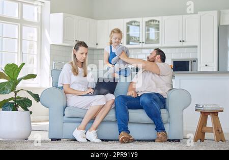 Caucasian mother working on a laptop and sitting on the couch while her daughter and husband play with a teddybear in the lounge at home. Woman using a laptop. Father and child playing with a toy. Stock Photo