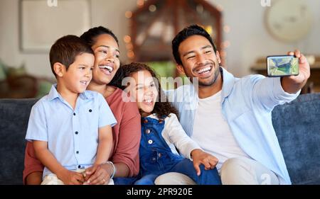 Family selfie on the sofa at home. Handsome young man of indian ethnicity taking pictures with his mixed race family on the sofa in the living room. Using his phone to take a picture for the album Stock Photo