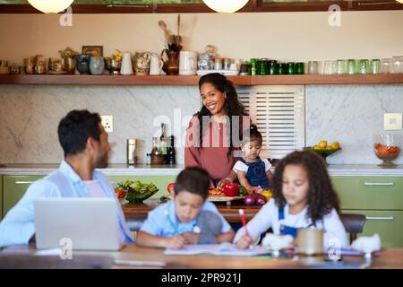 Happy mixed race mother cooking in the kitchen for her family. Dad working from home with laptop while kids do homework or being homeschooled. Family of five at home during coronavirus pandemic Stock Photo