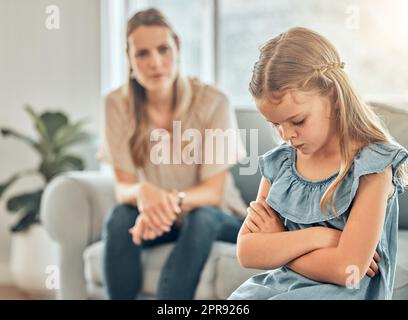 Sad little caucasian girl sitting in a living room with her arms crossed and feeling depressed while her mother watches annoyed. Young cute daughter feeling upset after disappointing a parent at home Stock Photo
