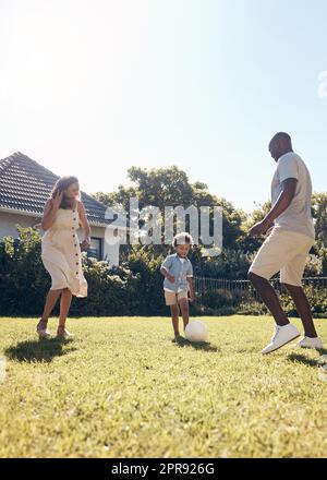 Happy mixed race family playing soccer outside in the garden at home. Parents enjoying kicking a ball with their son outside in the yard. Family bonding, having fun, playing together. Stock Photo